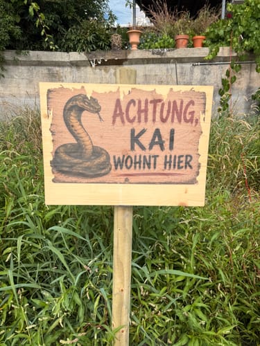 Reviewer's Holzschild bedruckt mit einer Kobra und dem Text Achtung, Kai wohnt hier, montiert an einem Pfosten im Garten.