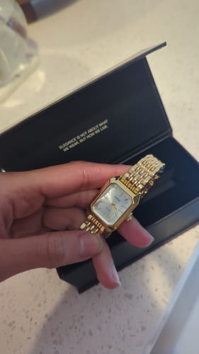 Reviewer's elegant gold Claire De Mains watch held in hand above its black branded display box.