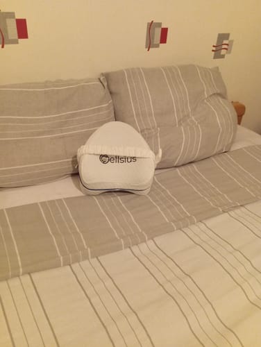Customer's Coussin genoux Cellsius blanc posé sur un lit avec des draps rayés gris et blancs.