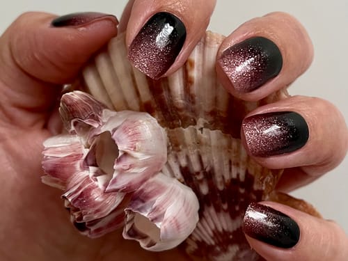 Customer's hand holding a seashell, showcasing Blush Nightfall UV gel nail wraps with a black to pink glitter gradient.