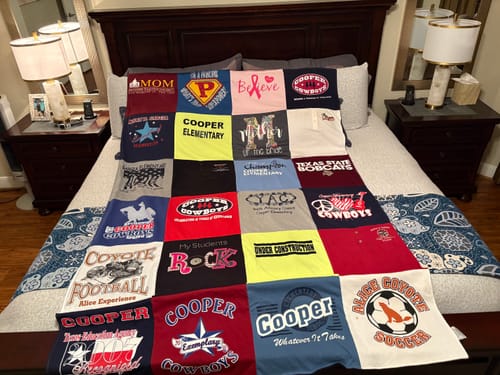 Customer's Project Repat T-Shirt Quilt spread across a bed, showing a patchwork of colorful school and sports shirts.