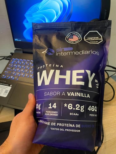 Customer's Proteína Whey sabor vainilla de Sin Intermediarios sostenida en una mano frente a una laptop.