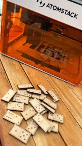 Customer's AtomStack P1 laser engraver with orange casing, surrounded by multiple wooden dominoes on a table.