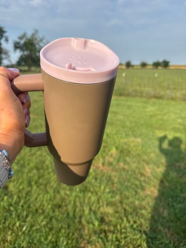 Customer's brown Thicc Buddy 40oz with a light pink lid held in a hand outdoors against a green field.