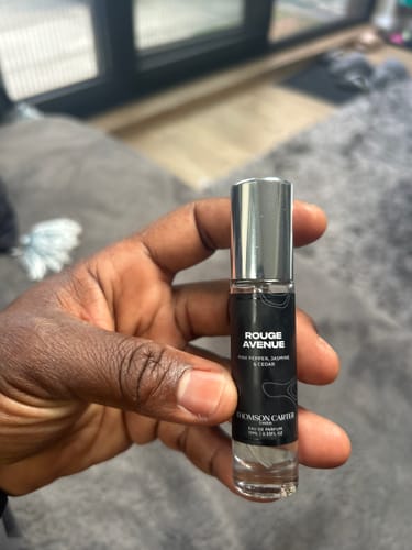 Reviewer's hand holding a small, cylindrical Rouge Avenue perfume bottle with a clear body and black label.
