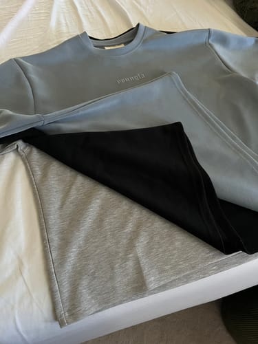 Customer's stack of Soft AF Tees in blue, black, and grey laid out flat on a bed.
