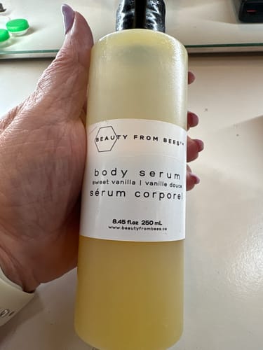 Customer's hand holding a bottle of Beauty From Bees Body Serum, Sweet Vanilla scent, in a clear plastic pump bottle.