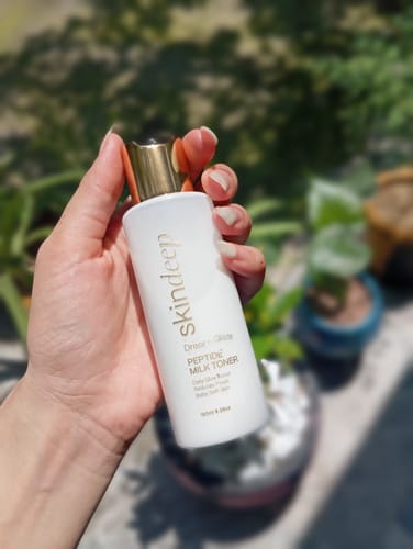 Reviewer's hand holding a white bottle of Dream Glaze Peptide Milk Toner against a blurry outdoor background.