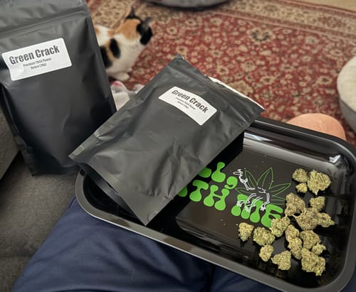 Customer's Green Crack flower in black pouches and on a graphic rolling tray with a cat in the background.
