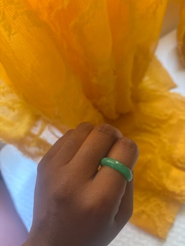 Customer's hand wearing a smooth, green jade ring, set against a soft yellow fabric background.