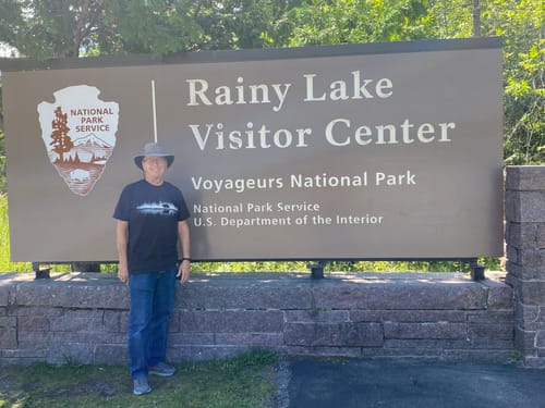 Customer's Bear Reflection T-Shirt, black color, worn while standing at the Rainy Lake Visitor Center sign.
