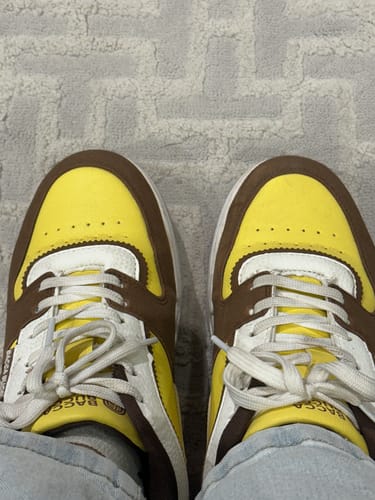 Reviewer's pair of FLORA sneakers in yellow and brown worn on a carpeted floor.