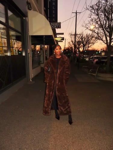 Customer's brown Lucie Faux Fur Maxi Coat worn by a person on a city sidewalk at sunset.