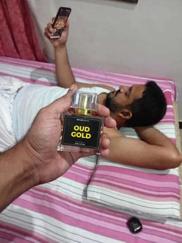 Customer's hand holding a bottle of The Royal Oud Collection Oud Gold perfume with a person relaxing on a bed behind.