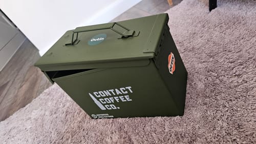 Customer's olive green M2A1 Ammo Tin with Contact Coffee Co. branding sitting on a textured carpet.