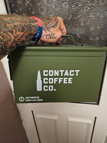 Customer's hand holding the green steel Coffee Resupply ammo tin with the Contact Coffee Co logo.