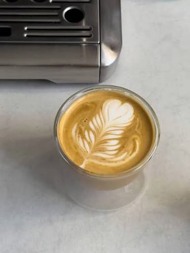 Customer's FLUR Latte Glass filled with coffee featuring heart-shaped latte art, placed next to a metal espresso machine.