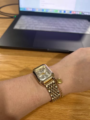 Customer's gold Claire De Mains watch on their wrist with a delicate bracelet against a blurry laptop background.