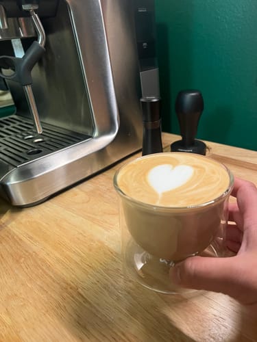 Reviewer's hand holding a FLUR Latte Lovers glass filled with coffee featuring heart-shaped latte art.