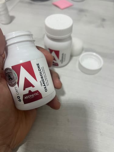 Customer's hand holding a white bottle of Aesthetic Labs Oxandrolona, close-up with another bottle in the background.