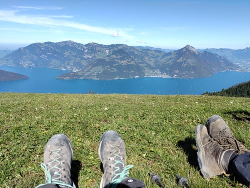 Customer's hiking boots resting on grass with a scenic mountain and lake view, made possible by Bosvera Plus Kapseln.