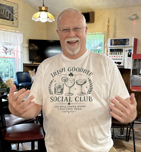 Customer's Irish Goodbye Club T-Shirt featuring a cocktail graphic, worn by a smiling person in a pub.