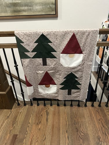 Customer's Gnomes Gone Wild quilt top hanging on a wooden banister, displaying gnome and pine tree patterns.