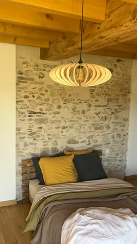 Customer's Mini NEO suspension lamp in wooden slats hanging in a bedroom with a stone wall and wooden ceiling.