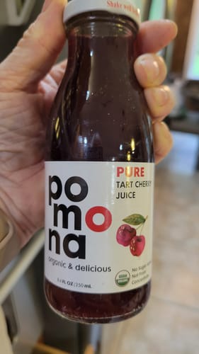Customer's hand holding a glass bottle of Pomona Organic Tart Cherry Juice with a clear label.