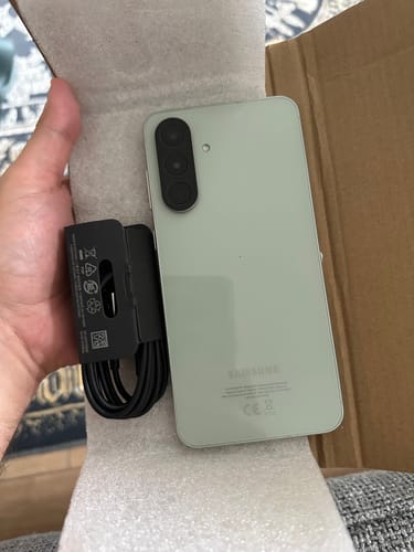 Customer's light green Samsung Galaxy A56 smartphone held in a hand next to a USB charging cable inside a box.