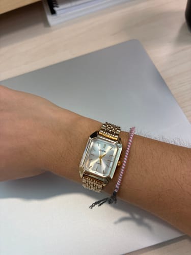 Customer's gold Claire De Mains watch with a rectangular face on a wrist next to a thin beaded bracelet.