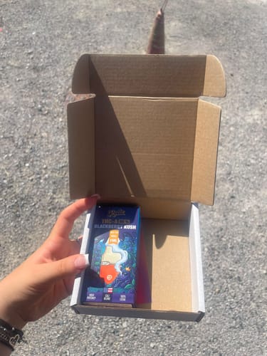 Reviewer's hand holding an open brown shipping box containing a Blackberry Kush THC-A vape cartridge package.