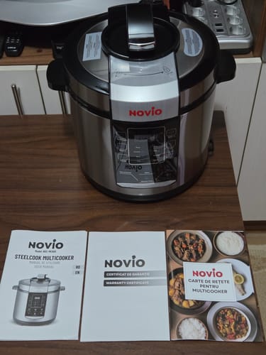 Customer's Novio SteelCook multicooker placed on a table next to its user manual, warranty, and recipe book.