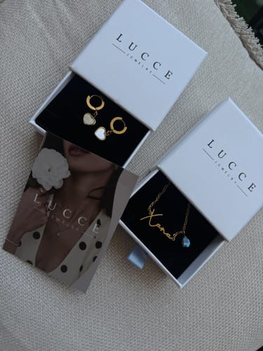 Customer's Lucce Signature Necklace, gold name script with teardrop stone, inside a box next to gold heart earrings.