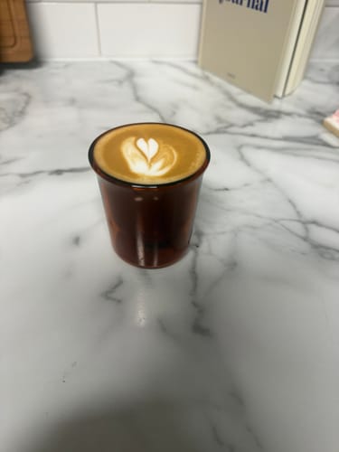Customer's amber-colored FLUR Cortado Glass filled with coffee featuring heart-shaped latte art, set on a marble counter.