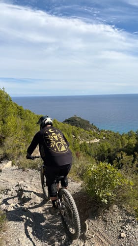 Reviewer wearing a black Occult MTB jersey riding a mountain bike on a coastal cliff trail.