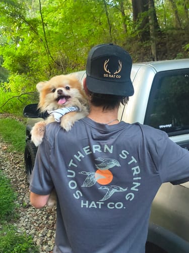 Customer's grey Shotgun Sunrise Tee worn by a person holding a small dog near a truck.