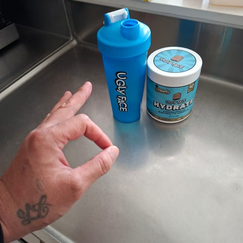 Customer's hand making an okay gesture next to a blue UGLYFACE HYDRATE shaker bottle and a tub of the product.