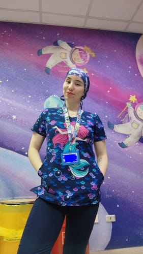 Reviewer wearing a Cherokee Disney TF626 top with a colorful mermaid print while standing in a space-themed room.