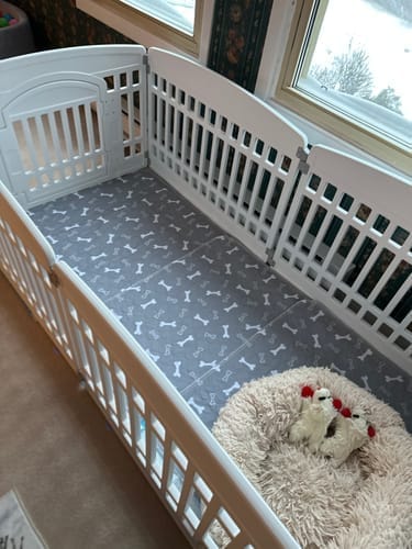 Tapete SniffPad cubriendo el piso de un corral blanco para cachorros, con una cama de peluche