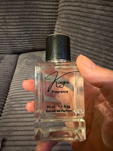 Kundenhand hält Kiage 286 Parfümflakon in einer transparenten, rechteckigen Glasflasche vor einem grauen Stoffhintergrund.