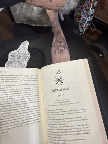 Reviewer reading a book while a fresh floral tattoo is on their shin, using the Signature Tattoo Numbing Cream