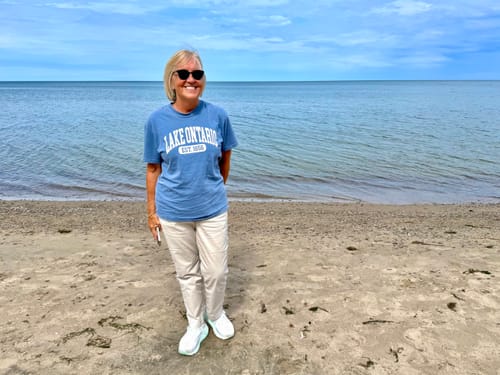 Customer's blue Lake Ontario Varsity T-shirt worn by a person standing on a sandy beach.
