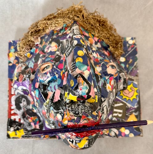 Customer's artistic collage craft shaped like a face with a straw-like top, featuring a purple pencil held in its mouth.