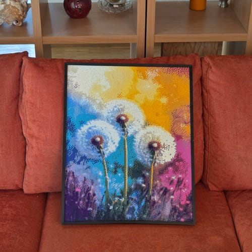 Kundenfoto eines eingerahmten Diamond Paintings mit Löwenzahn-Motiv in einem schwarzen Aluminium-Bilderrahmen auf einer Couch.