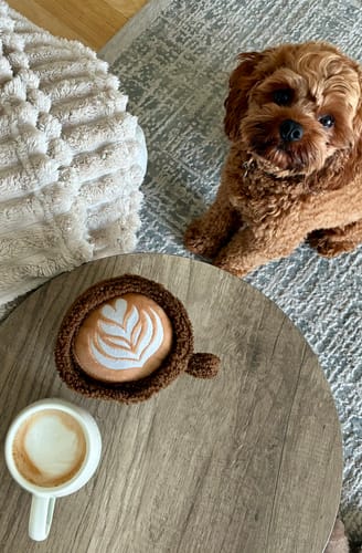 Customer's Flat White Cup Toy sitting on a wooden table next to a real coffee cup with a fluffy puppy watching.