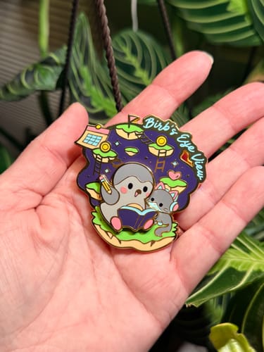 Customer's Birb's Eye View enamel pin, held in a hand over a green plant.