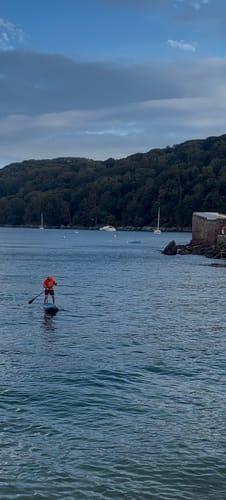 Customer's Aquaplanet PACE paddle board with a person in orange paddling on calm water near a wooded shoreline.