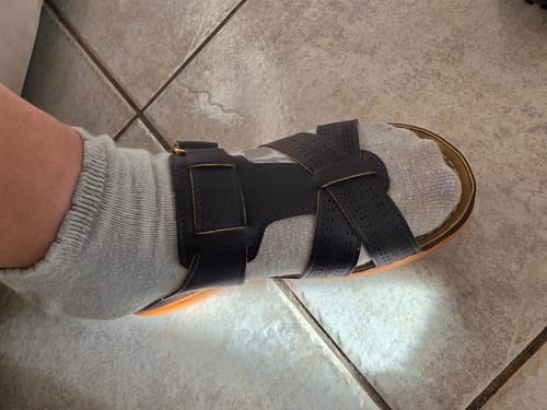 Customer's foot wearing a black, strapped orthopedic sandal over a light sock, placed on a tiled floor.