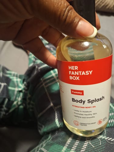 Customer's hand holding a clear bottle of Yummy Body Splash hydrating body oil over plaid pants.